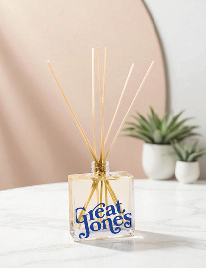 A clear glass reed diffuser with blue text and wooden reed sticks sits on a white surface, with small white potted succulents and a soft pink circular backdrop in a minimalist modern setting