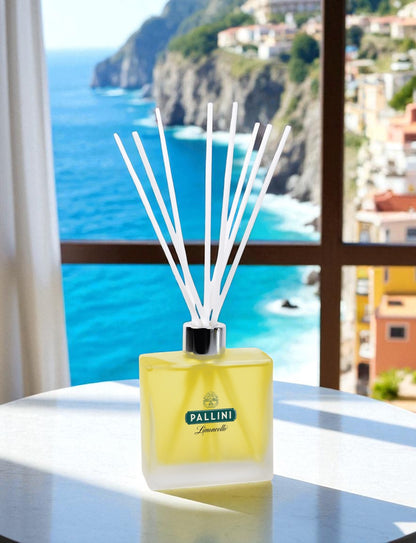 A frosted glass reed diffuser with tinted yellow fragrance; branding and white reed sticks sits on a white round table by a window, overlooking a stunning Mediterranean coastal view with turquoise waters, dramatic cliffs, and colorful hillside buildings