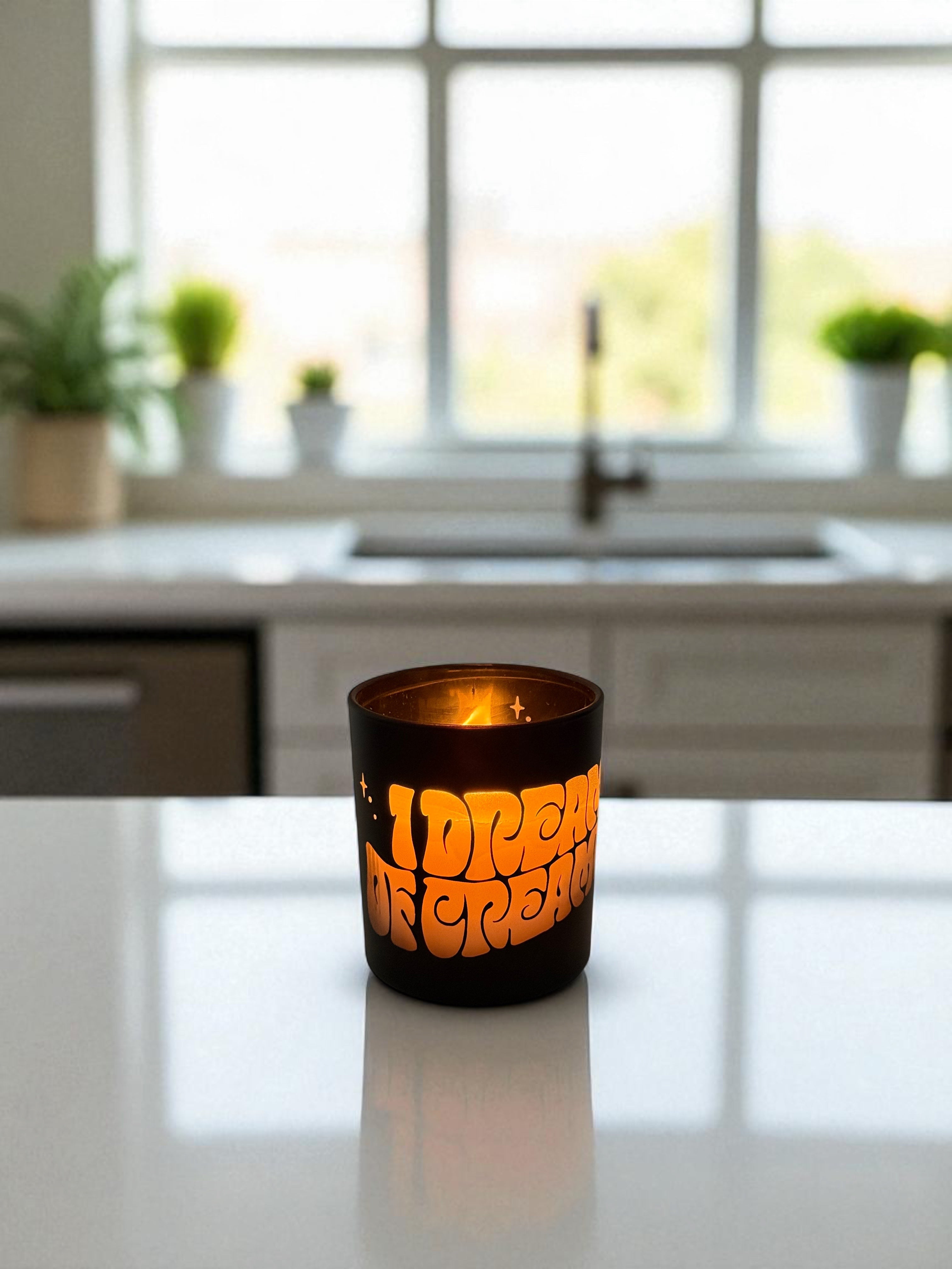 A lit 14 oz. black candle with engraved text sits on a white kitchen island countertop, with a modern kitchen featuring white cabinets, sink, and windows with potted plants in the background