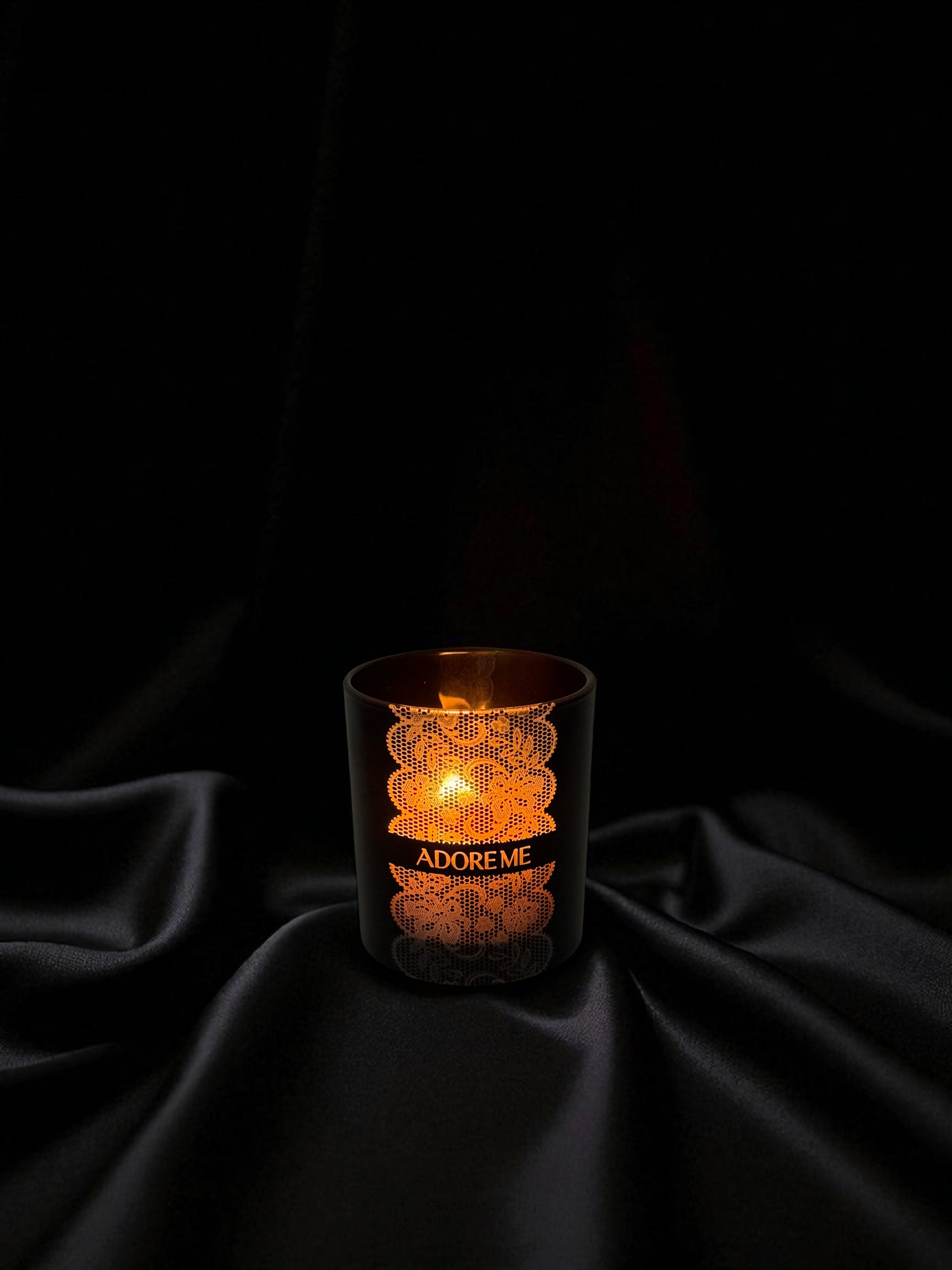 A 14 oz. black candle with wooden wick and engraved lace pattern sits a shiny black satin background 