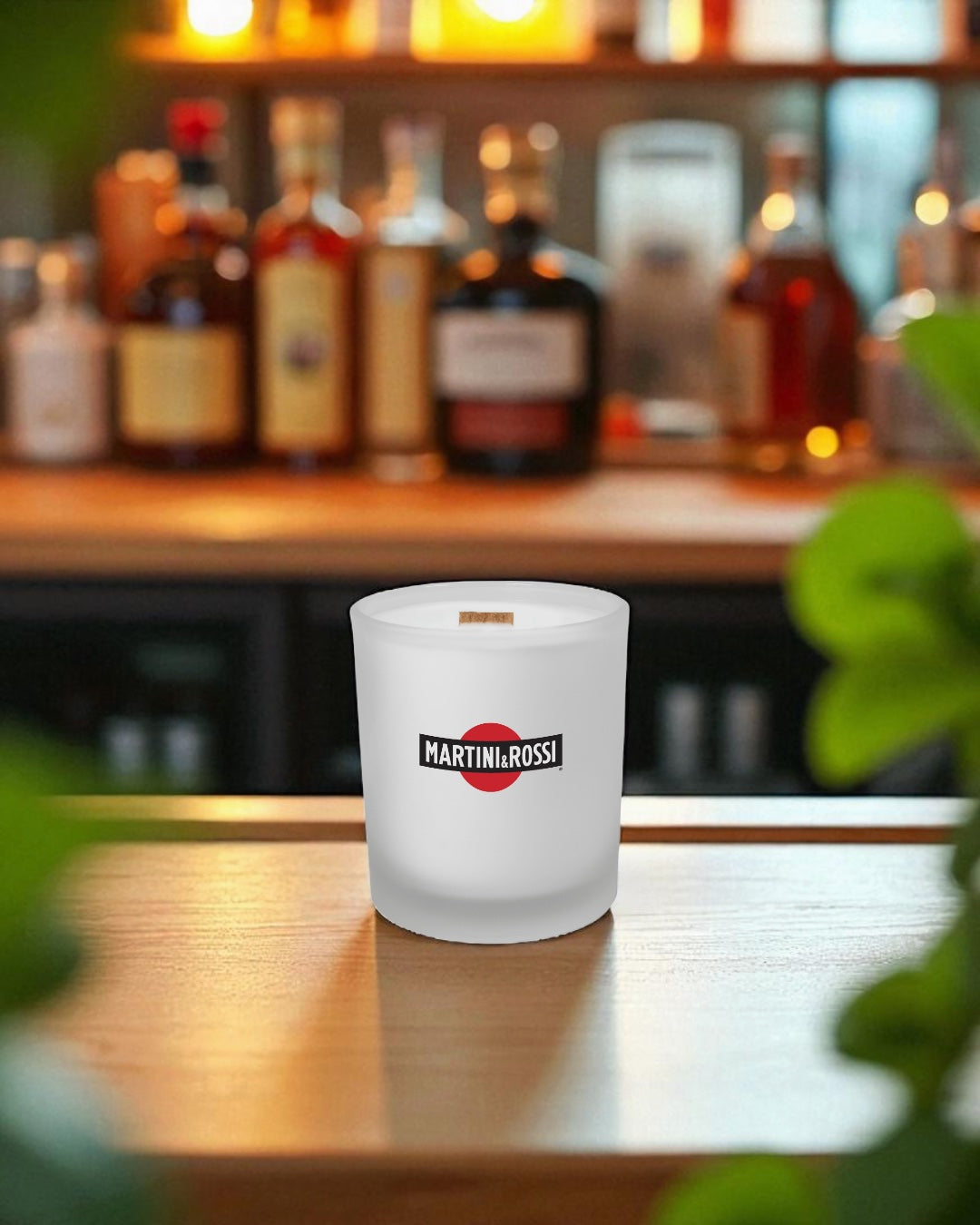 A frosted 14 oz. glass candle with wooden wick and logo sits on a polished wooden bar counter, with blurred liquor bottles and warm lighting in the background and green plant leaves visible in the foreground
