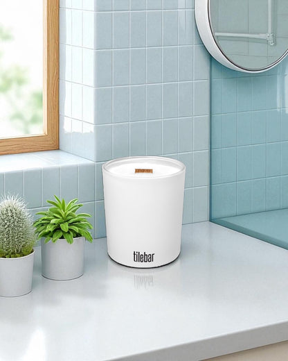 A white 14 oz. candle with wooden wick and black text sits on a white bathroom countertop next to small potted plants, with light blue subway tiles, a round mirror, and wooden window frame in the background