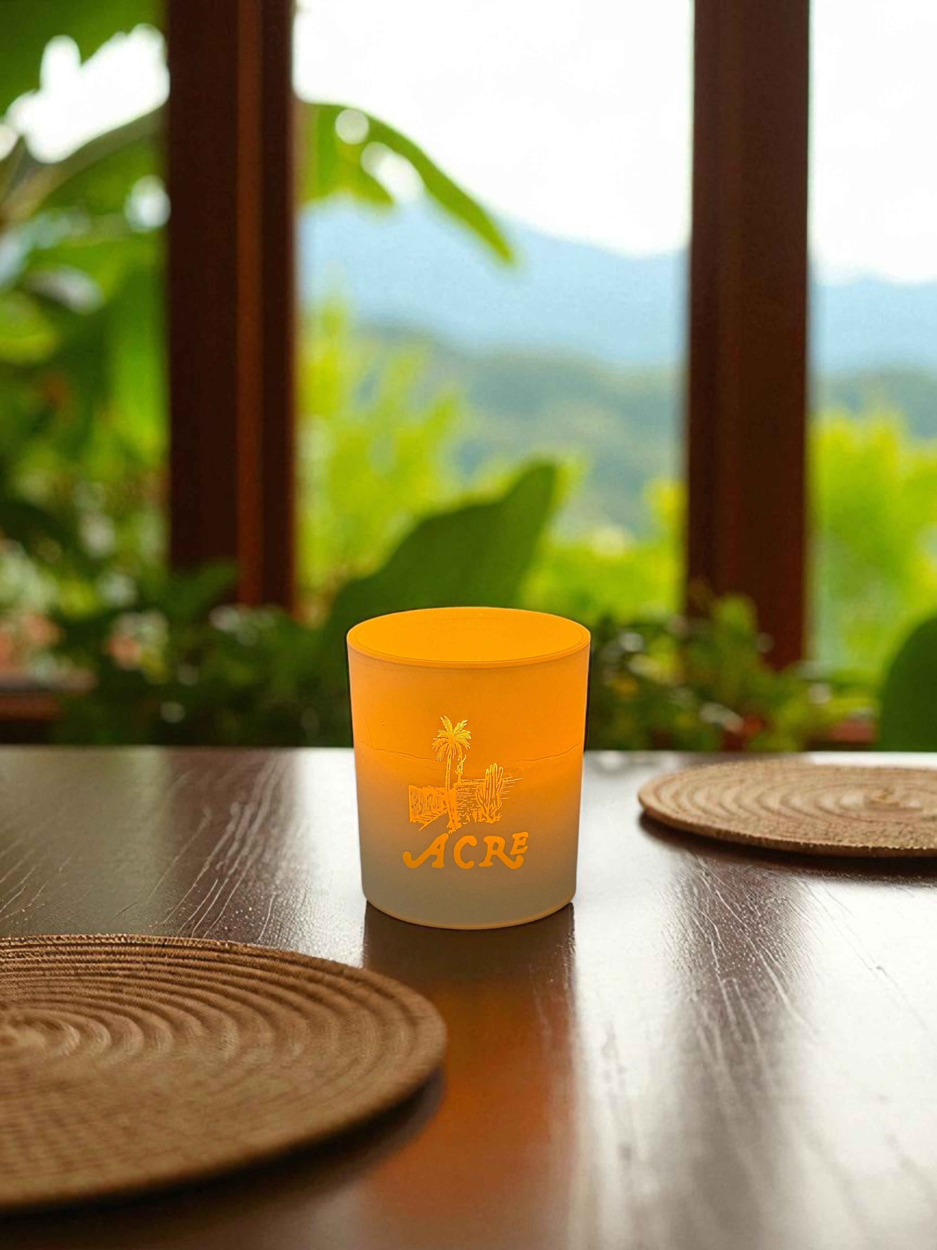 A 14 oz. white candle with engraved palm tree logo sits on a dark wooden dining table with woven placemats, overlooking a tropical view through large windows with lush green foliage