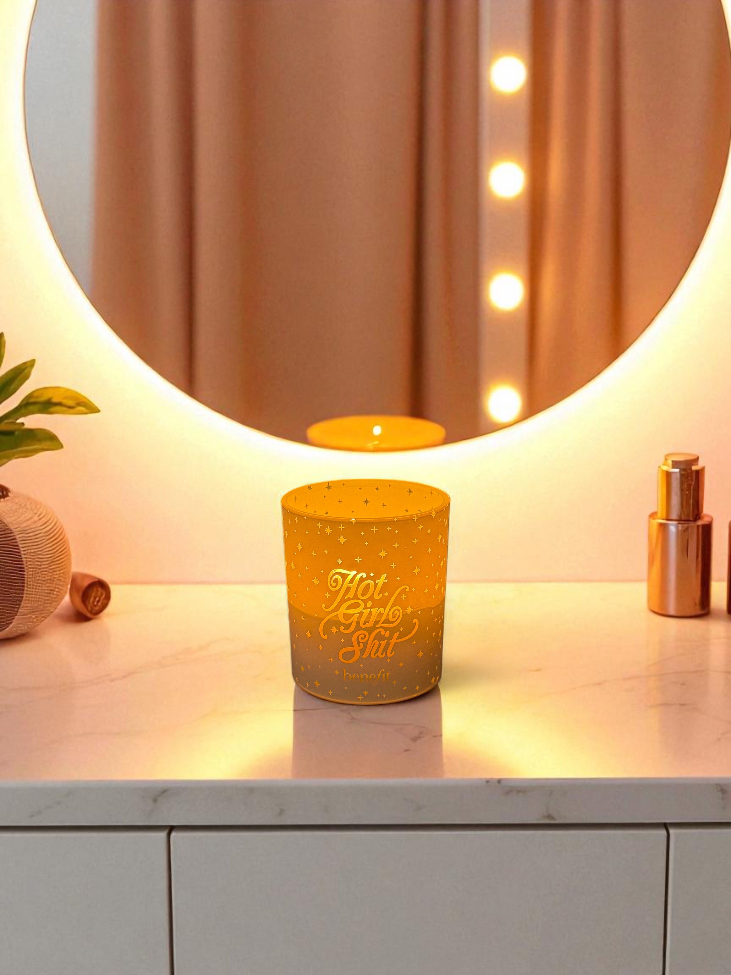 A lit 14 oz. white candle with engraved text and star pattern sits on a white marble vanity counter, with a round illuminated mirror, rose gold beauty products, and warm lighting in a modern bathroom setting