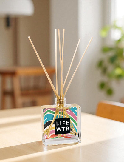 A clear glass reed diffuser with colorful pattern and text with wooden reed sticks sits on a wooden counter next to a plant at the distance
