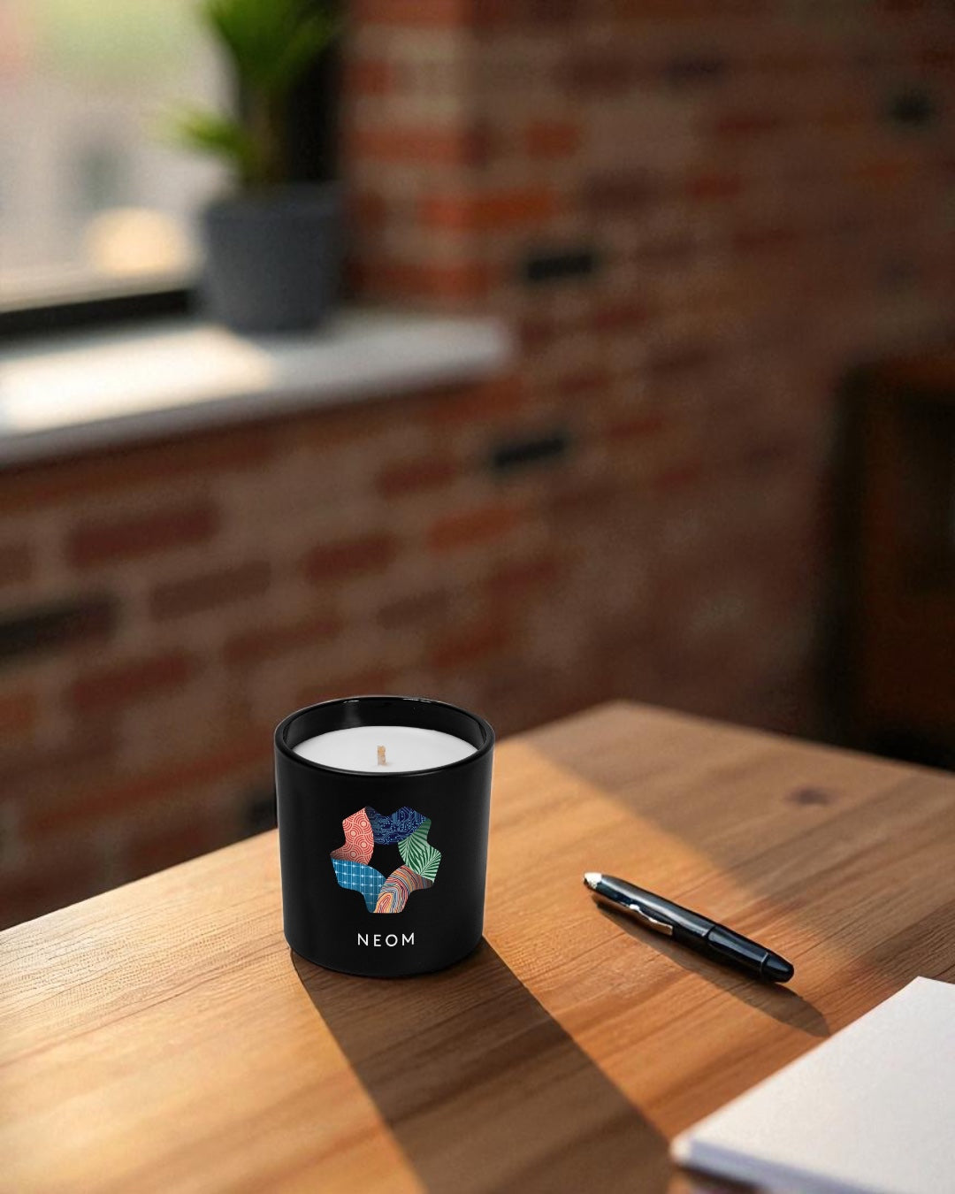 A black 9 oz. candle with a colorful geometric design sits on a wooden desk next to a pen and notebook. The candle has white wax visible at the top and is positioned in a modern workspace with exposed brick walls in the blurred background.
