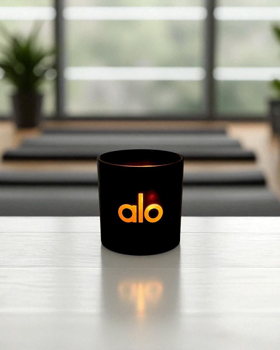A black 9oz. candle with engraved logo sits on a white reflective surface, with a warm glow visible inside. The candle is positioned in front of a blurred modern office setting with mats, plants, and large windows in the background.