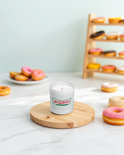A white 9 oz. candle with green and red logo sits on a round wooden board surrounded by colorful glazed donuts on a marble countertop, with a wooden donut display rack in the background
