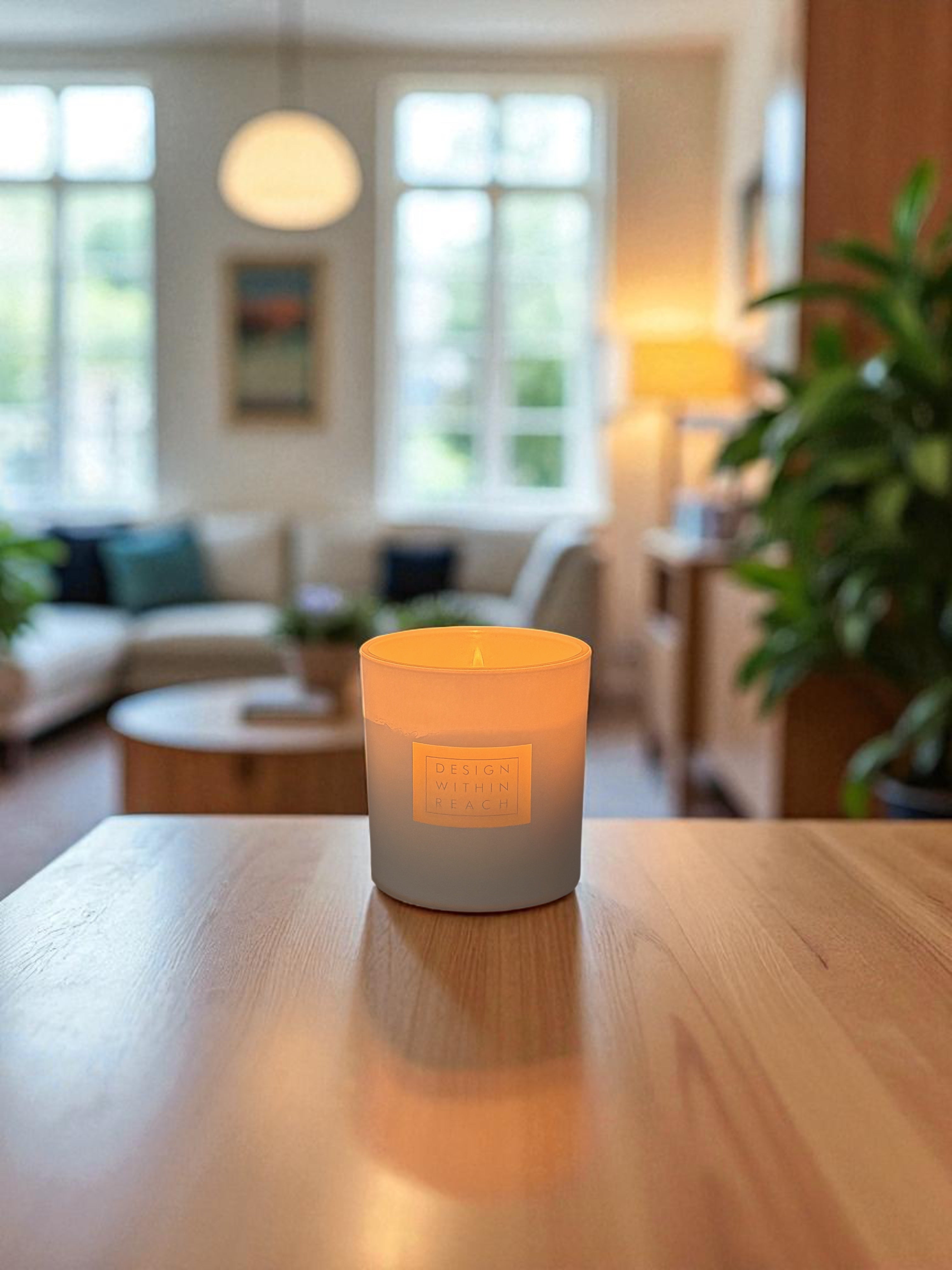 A white 9 oz. glass candle with engraved logo on the front sits lit on a wooden surface, casting a warm glow. The candle is positioned in an elegant interior space with classical artwork visible on the walls in the softly blurred background.