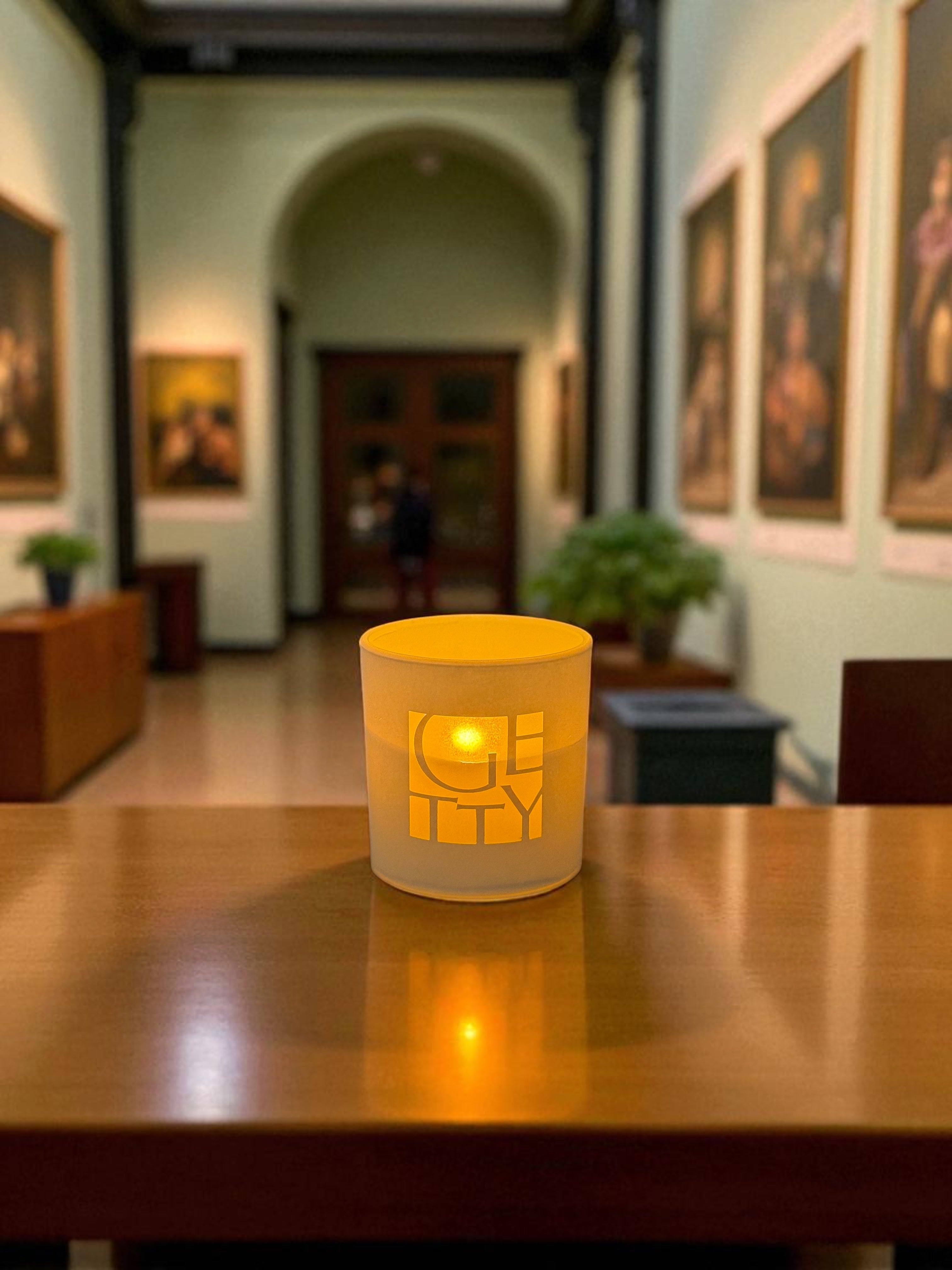 A white glass candle with logo etched on the front sits on a polished wooden table, glowing with warm amber light. The candle is positioned in an elegant museum gallery with classical paintings on white walls and an arched doorway in the background.